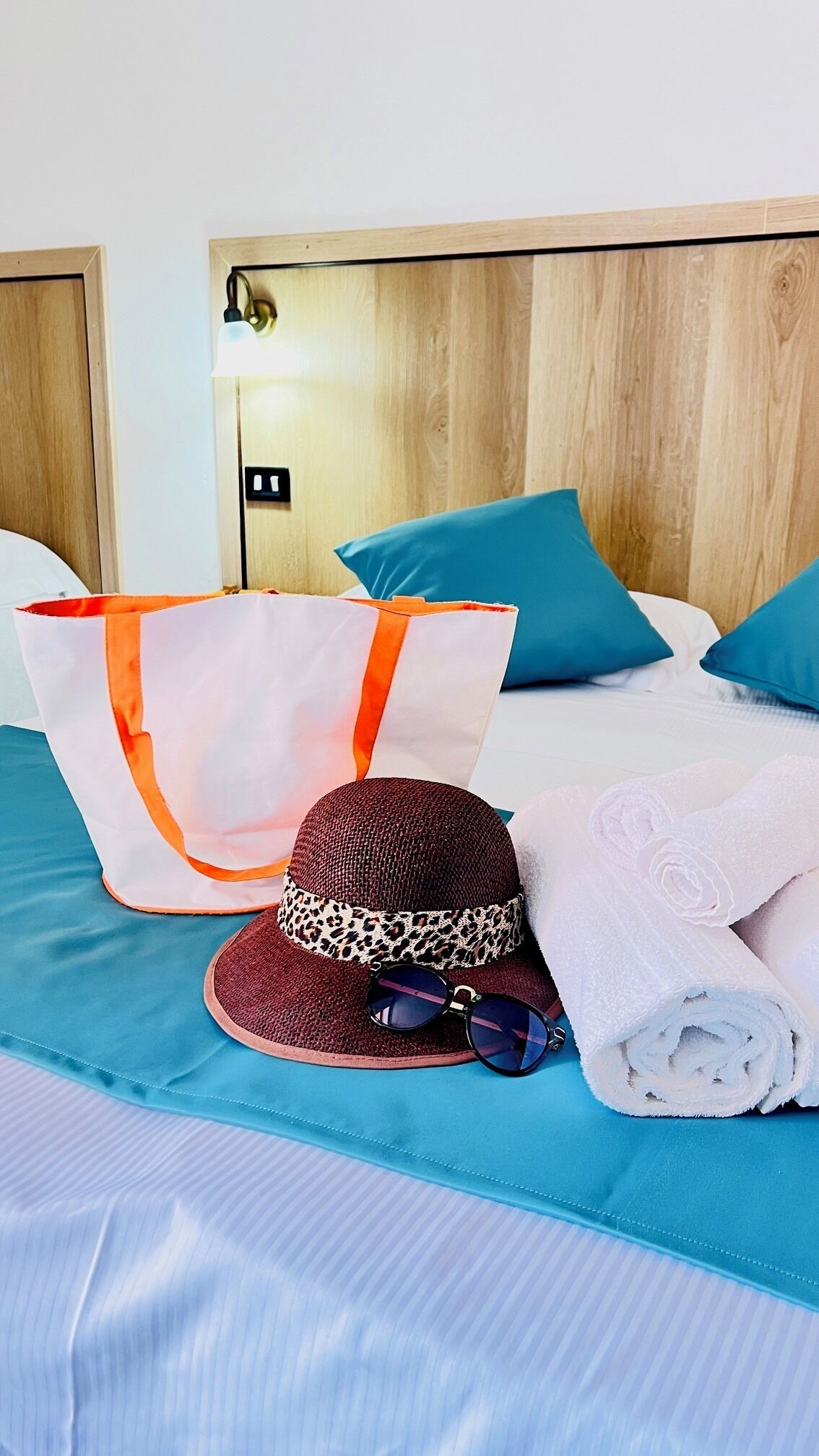 A bed with a blue bedspread, a white pillow, and a blue pillow. There is a white bag with orange handles on the bed.