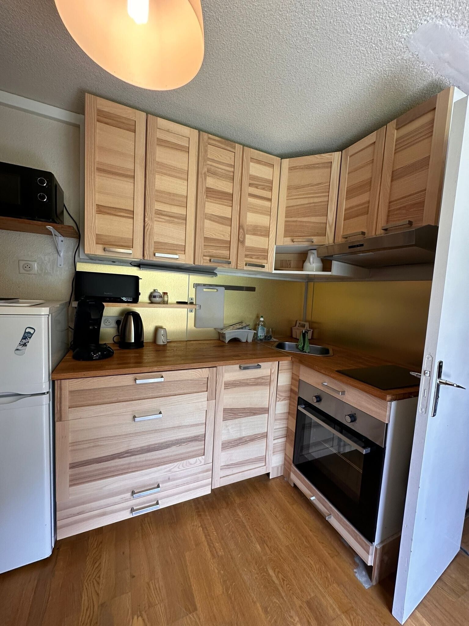 Een kleine keuken met houten kastjes, een magnetron, een koffiezetapparaat en een koelkast.