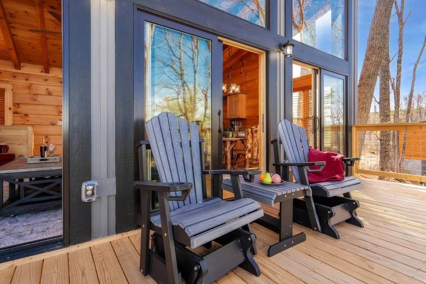 En træterrasse med to Adirondack-stole, et bord med frugt og udsigt til et interiør gennem store vinduer.