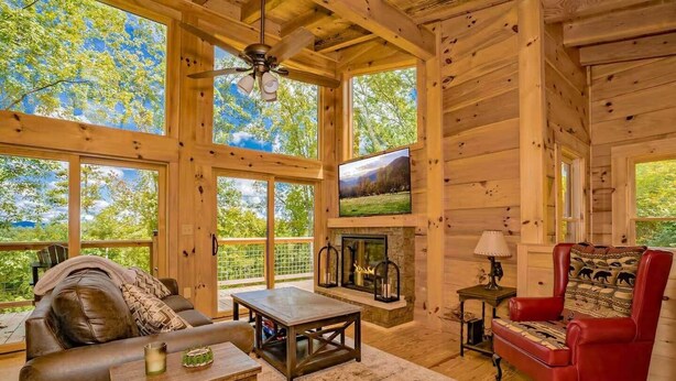 Interior acogedor de una cabaña de madera con chimenea, muebles de cuero y grandes ventanales que ofrecen vistas al exterior.