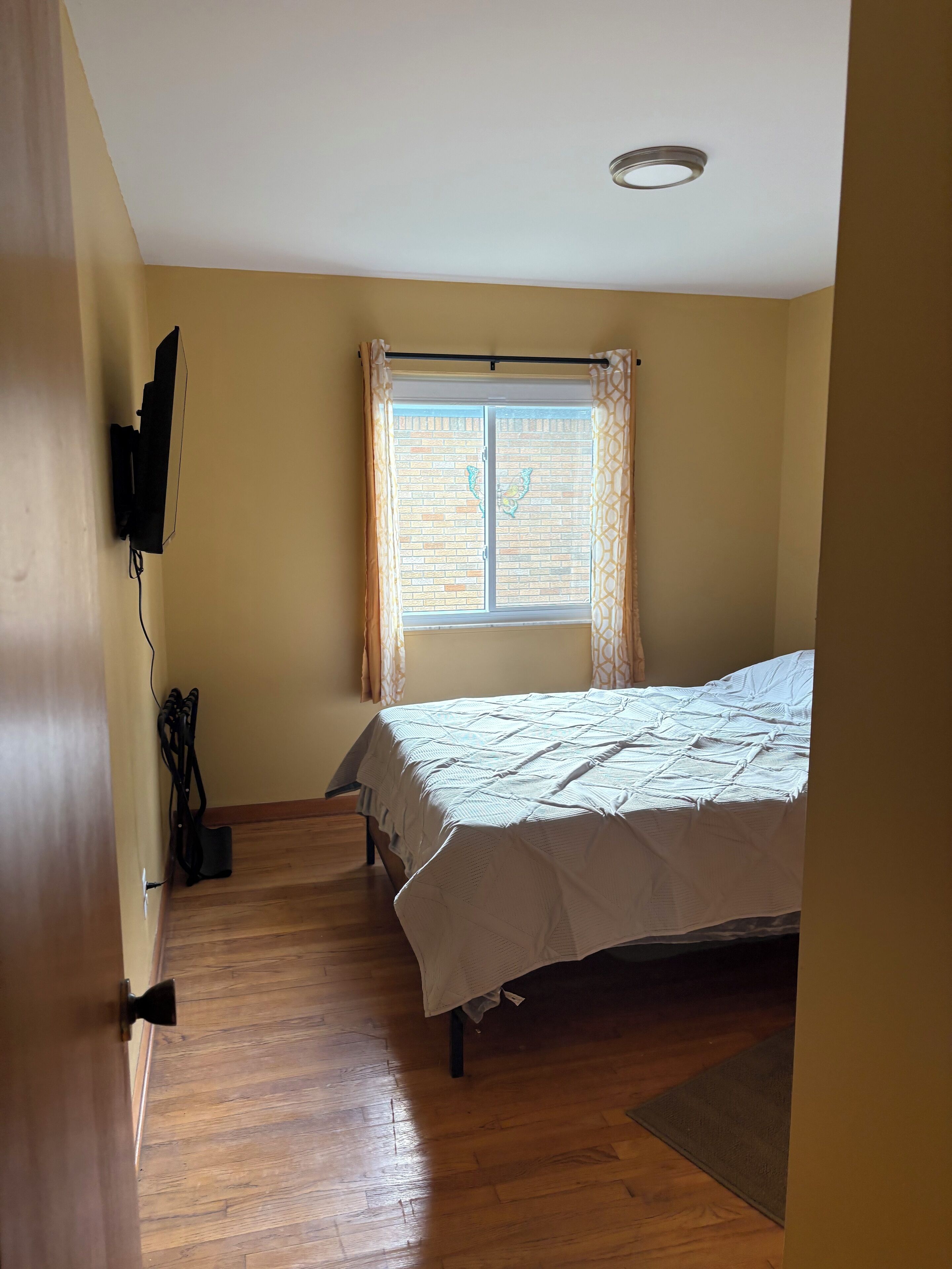 A bedroom with a bed, a window with curtains, a ceiling light, and a wooden floor.