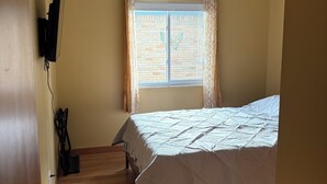 Une chambre à coucher avec un lit, une fenêtre avec des rideaux, un luminaire au plafond et un plancher en bois.