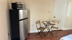 Une petite cuisine équipée avec un réfrigérateur et un micro-ondes en acier inoxydable, une petite table à manger avec deux chaises, et une porte blanche en arrière-plan.