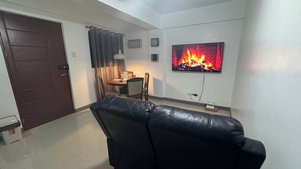 A living room with a black leather sofa, a dining table with chairs, a TV mounted on the wall displaying a fireplace, and a door with a handle. - Cozy Clean Space with Balcony - Near Entrance (Davao City)