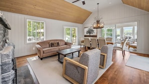 A spacious living room with a high ceiling, wooden beams, a stone fireplace, a large window, and a dining area with chairs and a table.
