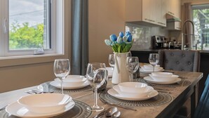 Een eettafel gedekt voor zes personen, met witte borden, glazen en bestek, een vaas met blauwe tulpen, en uitzicht op de keuken en het buitengebied.