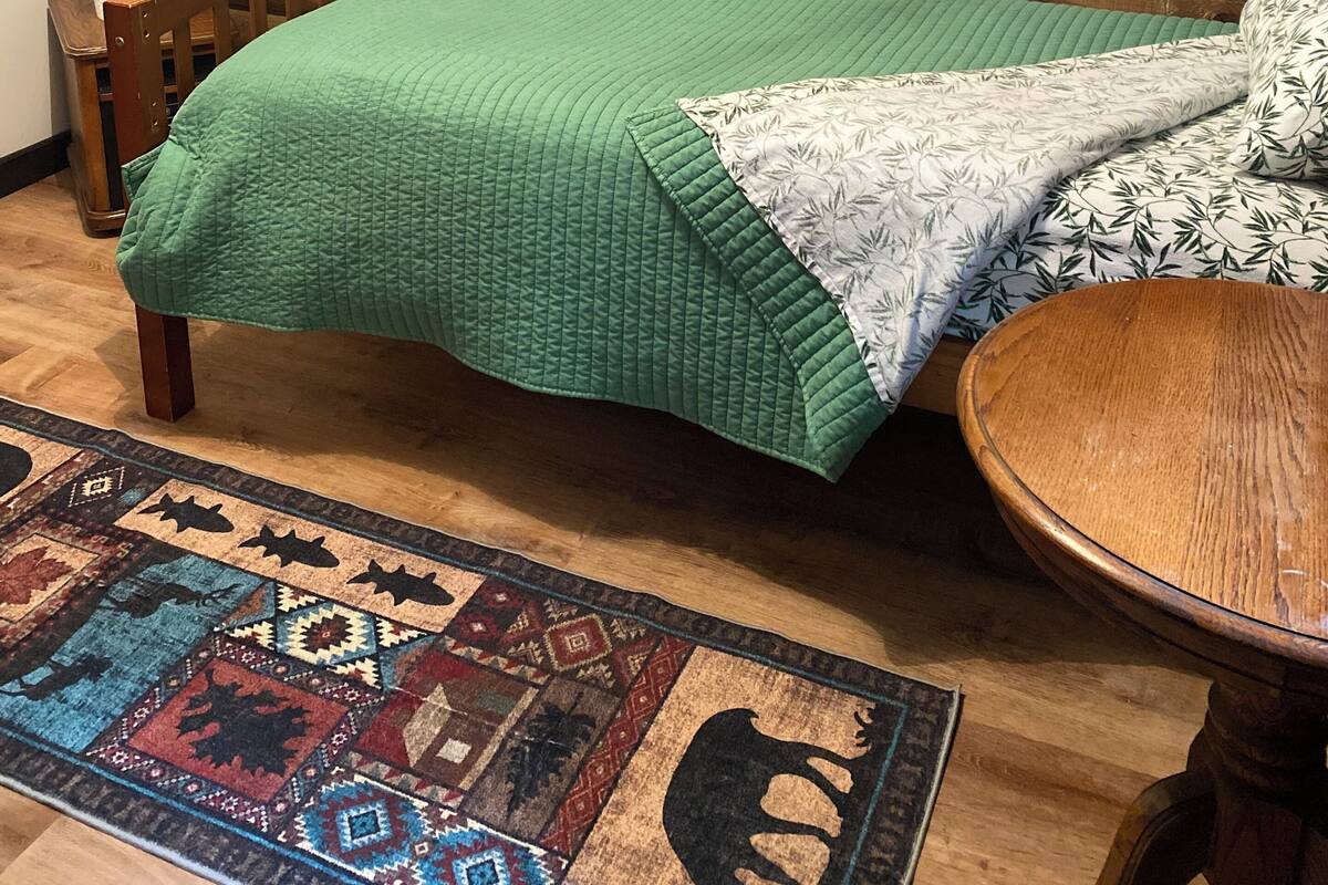 A log cabin room with a bed, a framed picture of trees and animals, a wooden chair, and a patterned rug.