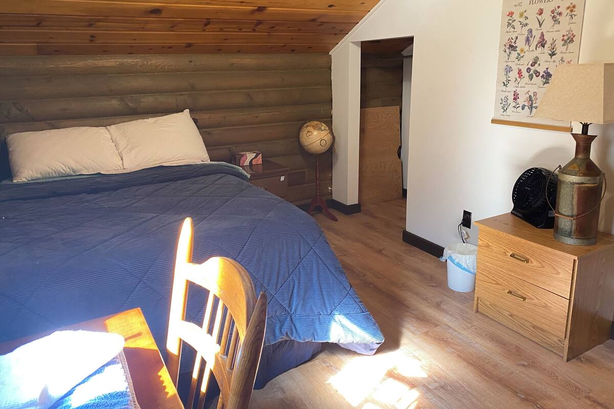 A cozy room with a wooden ceiling, a bed with a blue quilt, a wooden chair, a globe, a fan, and a vintage lamp.