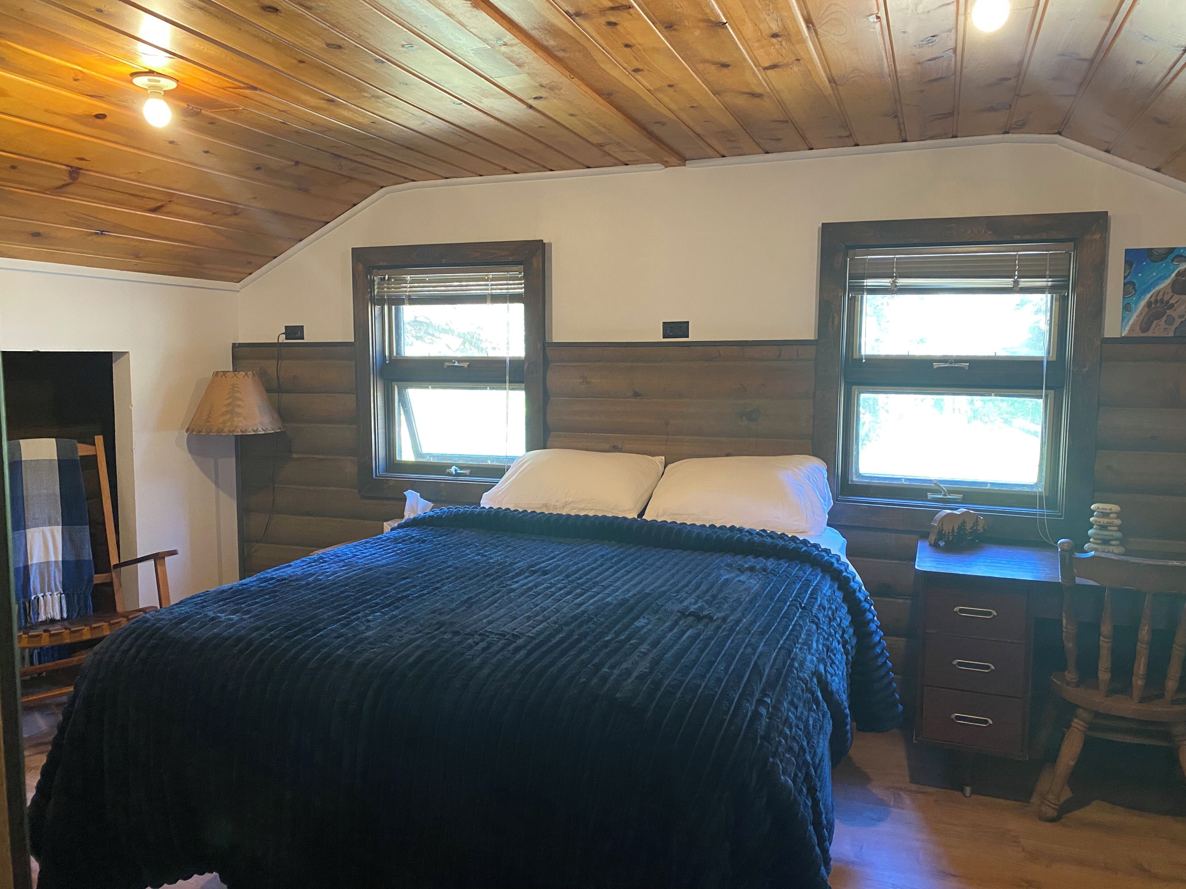 A bedroom with a wooden ceiling, a bed with a dark blanket, two windows with blinds, a nightstand, a dresser, and a lamp.