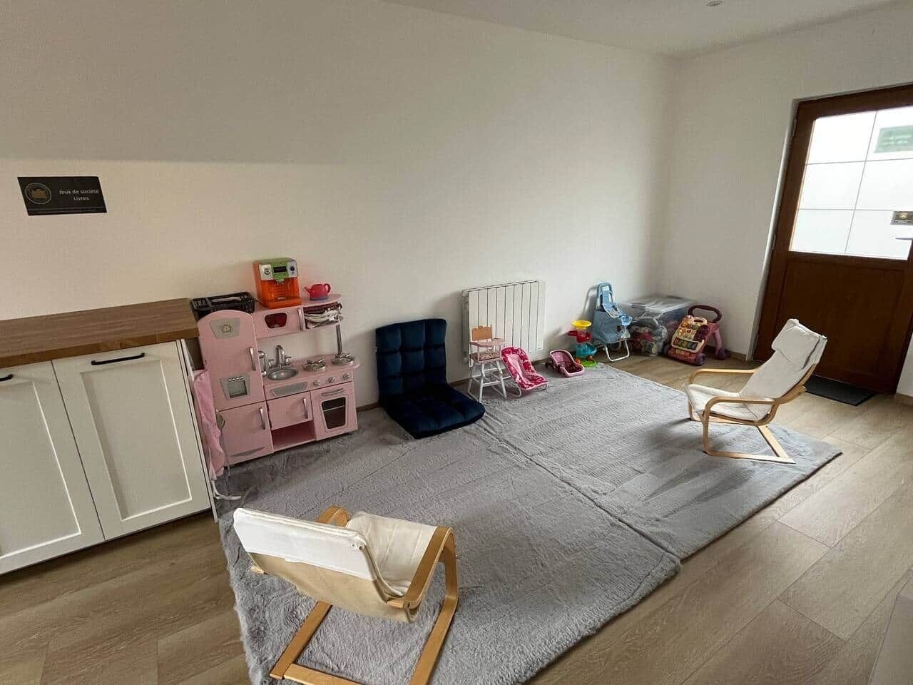 Une pièce avec un espace de jeux comprenant une cuisine miniature rose, un banc bleu rembourré et une chaise en bois.