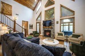 A spacious living room with a stone fireplace, wooden beams, and large windows. - Treat Yourself to a Stay at Sugar Plum Cottage (Broken Bow)