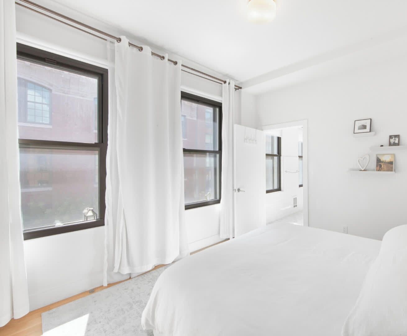 A modern bedroom with a large bed, two windows, a door, and a small shelf with decorative items.
