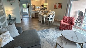 Un vaste salon comprenant un canapé d’angle gris, une table basse ronde et un fauteuil à motifs rouges. On aperçoit à l’arrière-plan un coin repas avec des chaises blanches et une cuisine aux meubles blancs.