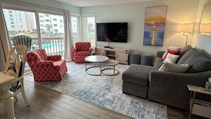 A modern living room with a flat-screen TV, a grey sofa, a red patterned armchair, a round coffee table, and a view of a building and a pool through large windows. - Cozy 1-bedroom, 1.5 bathroom condo in Emerald Isle with FANTASTIC ocean views! (Emerald Isle)