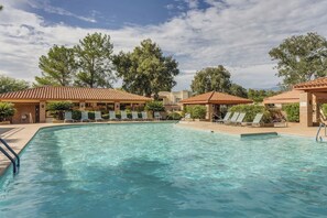 Una piscina con lettini e zone ombreggiate, circondata da alberi ed edifici.