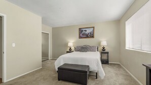 Une chambre à coucher avec un lit, deux tables de chevet avec des lampes, une commode, un banc, une porte et une fenêtre avec des stores.