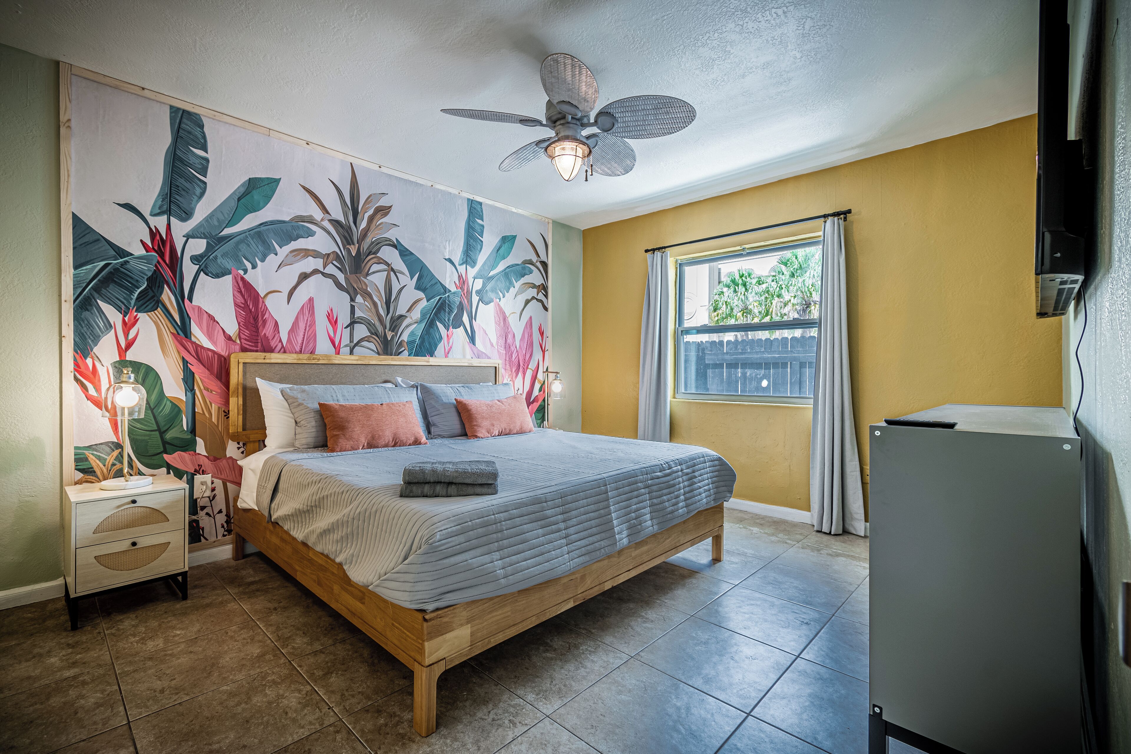 Quarto com cama, ventilador de teto, janela com cortinas e parede com um mural tropical.