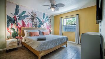 Quarto com cama, ventilador de teto, janela com cortinas e parede com um mural tropical.
