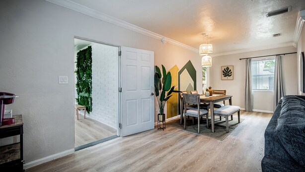 A modern living room with a dining area, featuring a wooden floor, a gray sofa, a dining table with chairs, and a wall-mounted plant. - 2/1 with private backyard in the heart of Ivanhoe (Orlando)