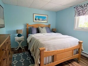 A bedroom with a wooden bed, a nightstand, a dresser, and a window with a view of greenery. - Soothing 4-bedroom house in welcoming Fryeburg perfect for families (Fryeburg)
