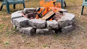 Ein Lagerfeuer mit brennenden Holzscheiten, umgeben von Steinen, zwei Adirondack-Stühle und ein grünes Haus mit einer Veranda im Hintergrund.