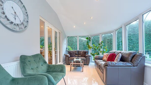 Un salón amplio con un sillón verde, un sofá gris con cojines rojos y una mesita de centro. Grandes ventanales ofrecen vistas al exterior, con vegetación.