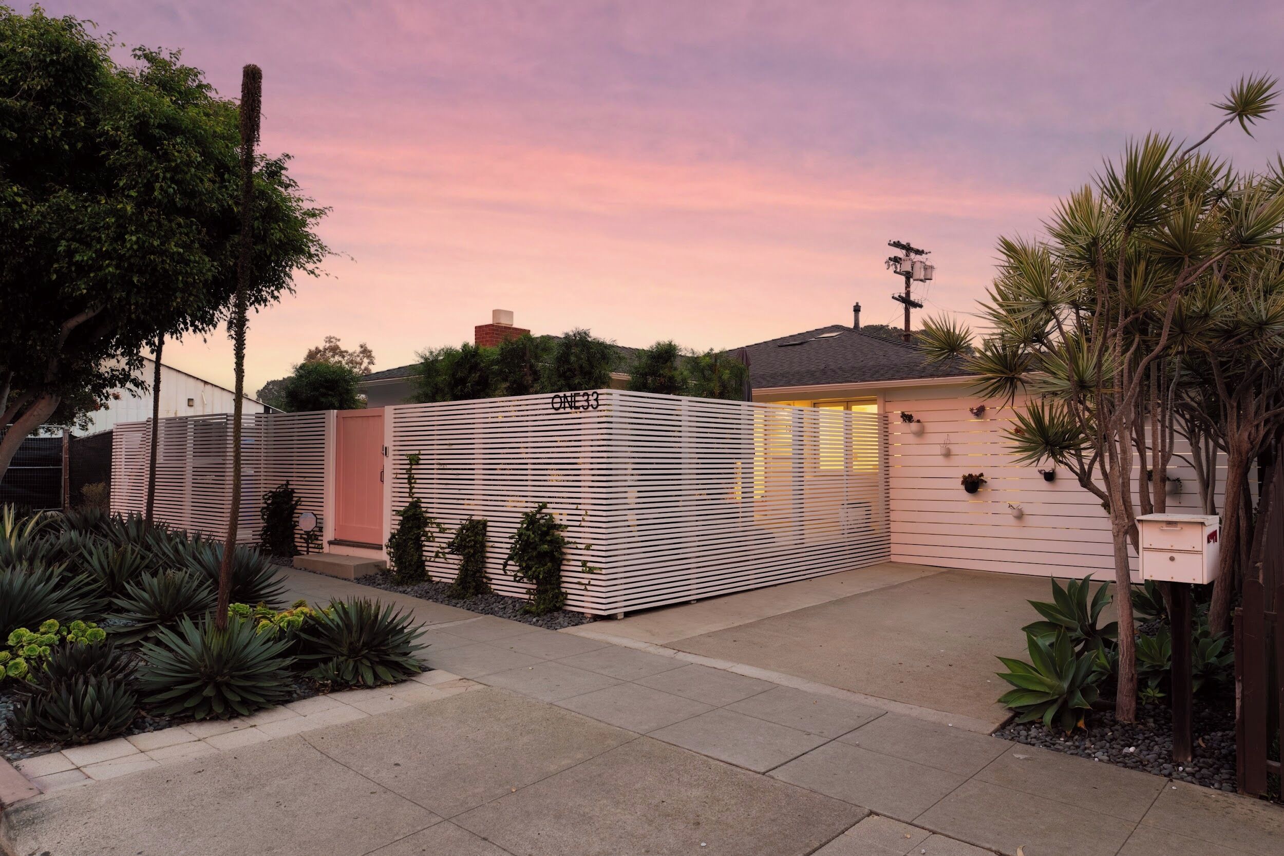 Een modern huis met een hek, een verzorgde tuin en een heldere hemel bij zonsondergang.