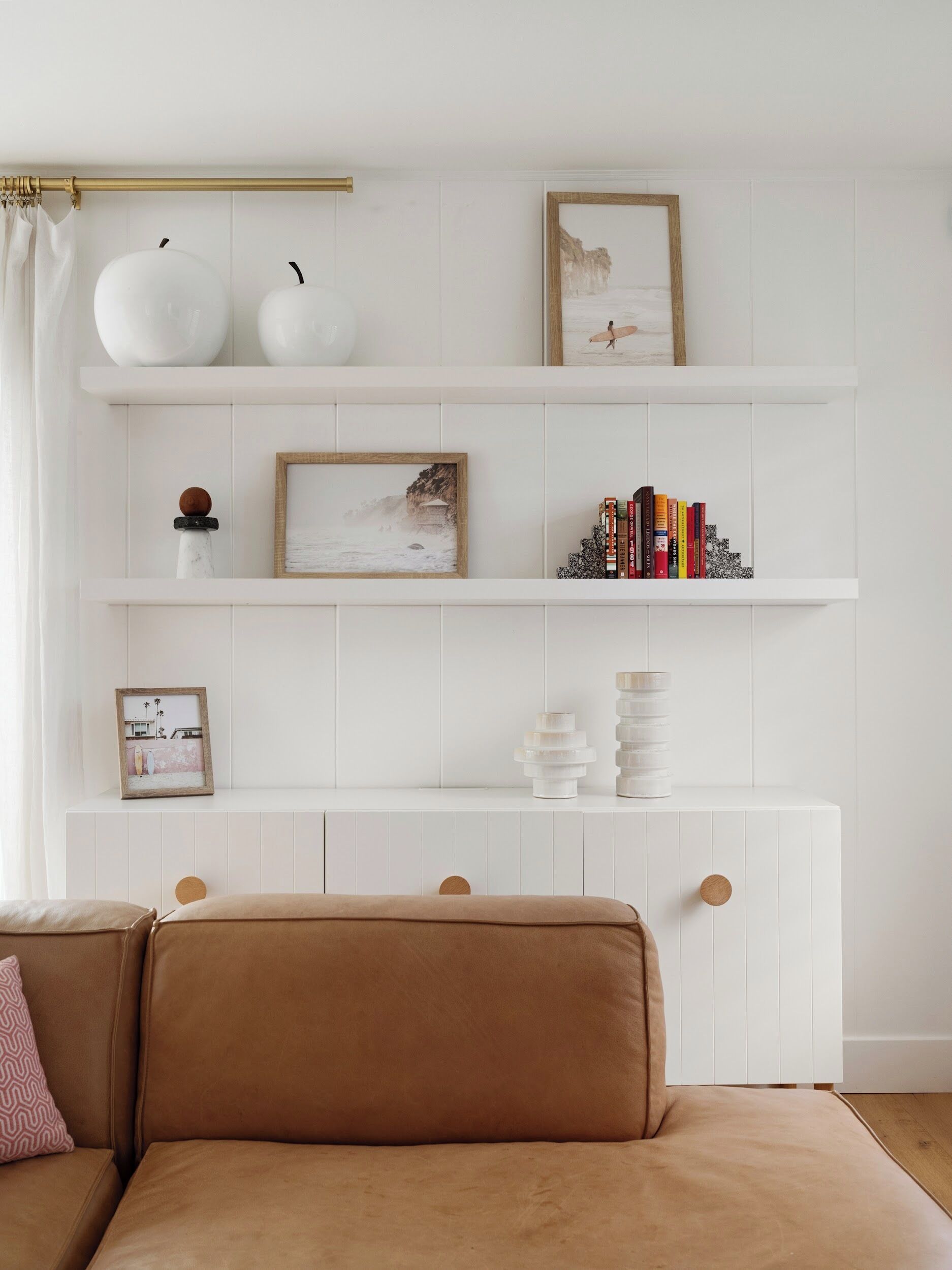 Una sala de estar con pared de azulejos blancos, un sofá marrón y un aparador blanco con cajones. Hay objetos decorativos en los estantes, incluyendo libros y cuadros enmarcados.
