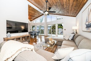 Un salón espacioso con ventilador de techo, televisor grande y vistas al exterior a través de amplios ventanales.
