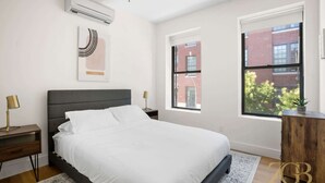 A modern bedroom with a large bed, two bedside tables with lamps, a wall-mounted air conditioner, and a window with a view of a brick building. - Renovated 2 Bed 2 Bath Apt in Prime Midtown - Elevator - Laundry (New York)