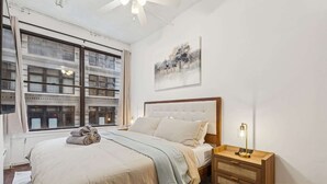 A bedroom with a large bed, a nightstand, a lamp, a window with a view of buildings, and a painting on the wall. - Spacious 3BR Flex 4 with Washer & Dryer in NoMad (New York)