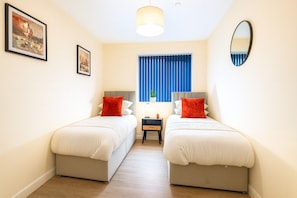 A room with two beds, a small table, a lamp, a window with blue curtains, and a mirror on the wall. - Cardiff 3BR Apartment| Contractors | Sleeps 8 (Cardiff)