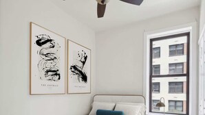 Un dormitorio con una cama, un ventilador de techo, una ventana y dos cuadros abstractos en la pared.