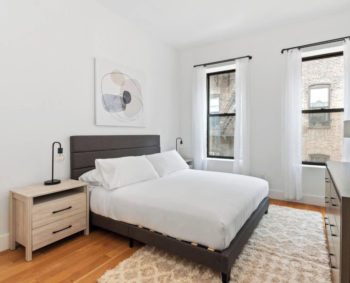 A modern bedroom with a bed, bedside tables, a nightstand, a large window with curtains, and a view of a brick building.