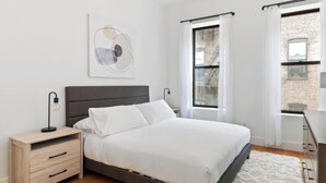 A modern bedroom with a bed, bedside tables, a nightstand, a large window with curtains, and a view of a brick building. - Gorgeous and Fully Equipped Luxury 1 Bedroom Apt! (New York)
