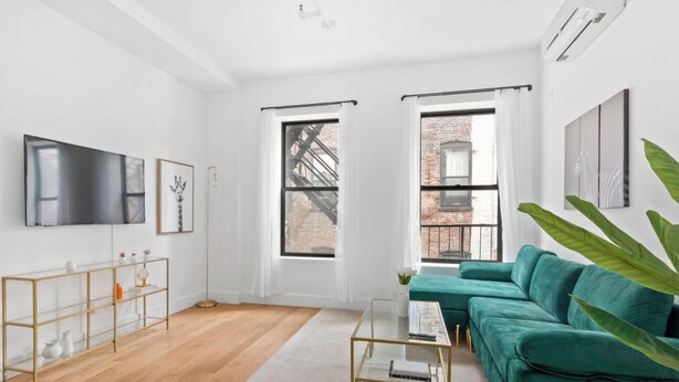 A modern living room with a teal sofa, a glass coffee table, and a flat-screen TV mounted on the wall. There are two windows with white curtains, and a large green plant is visible. - Gorgeous and Fully Equipped Luxury 1 Bedroom Apt! (New York)