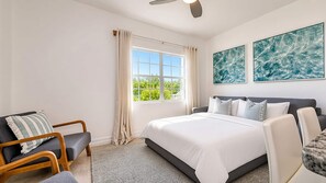 Une chambre à coucher comprenant un lit, deux chaises, un ventilateur de plafond, une fenêtre avec des rideaux et deux tableaux abstraits accrochés au mur.