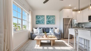 Un salon moderne avec un canapé gris, une table basse en bois et un coin cuisine doté de meubles blancs et d’un réfrigérateur en acier inoxydable.