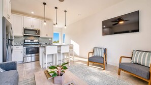 Una cucina moderna con elettrodomestici in acciaio inossidabile, una zona pranzo con sedie bianche e un soggiorno con divano e poltrona.
