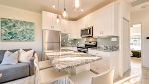 A modern kitchen with white cabinets, a granite countertop, stainless steel appliances, and pendant lighting. - Beach Paradise 1 Bedroom with Pull Out Sofa (West Bay)