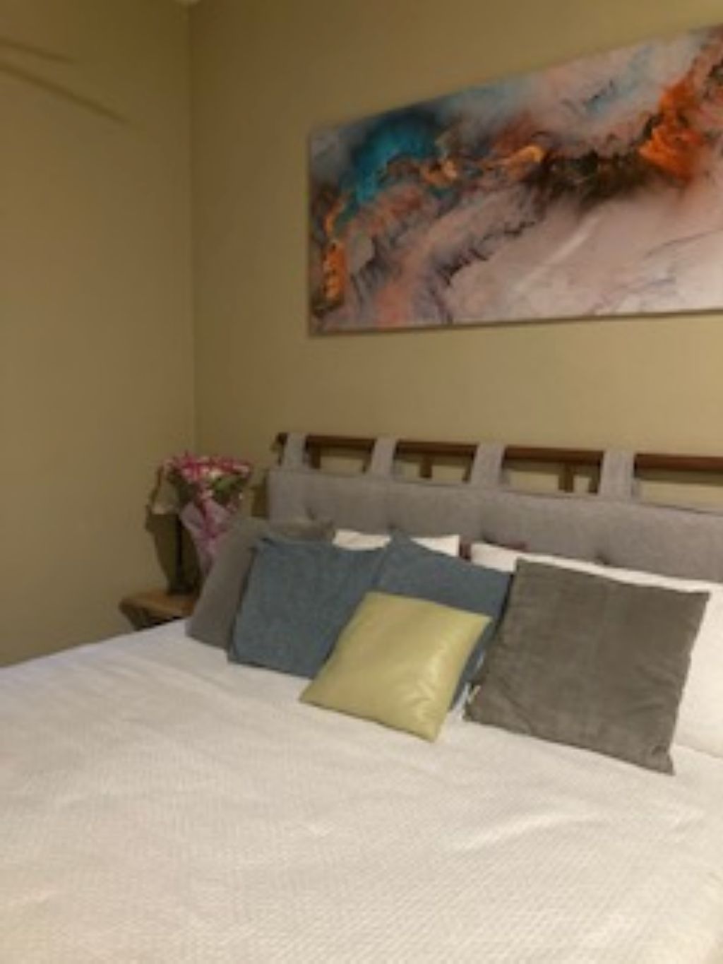 A bed with multiple pillows and a headboard, a painting on the wall, and a nightstand with a vase of flowers.