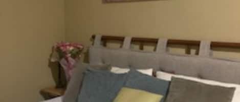 A bed with multiple pillows and a headboard, a painting on the wall, and a nightstand with a vase of flowers.