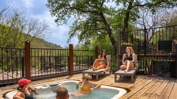 A hot tub surrounded by lounge chairs on a wooden deck with a metal fence and trees in the background. - Guadalupe Riverfront | Private Hot Tub | Sleeps 14 (New Braunfels)