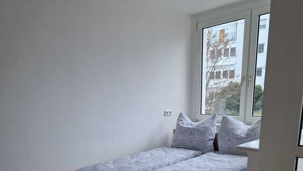 Ein kleines Zimmer mit einem Einzelbett, Holzboden, einem Fenster mit Blick auf Gebäude und einer weißen Wand.