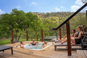 A wooden deck with a hot tub, lounge chairs, and a view of a hilly landscape. - Historic Guadalupe Riverfront Home | Sleeps 10 | Private Hot Tub + Billiards Room (New Braunfels)