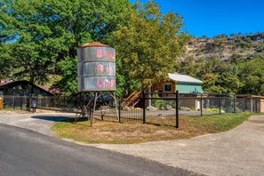 Ein rustikaler Wasserturm mit der Aufschrift 'BILL GOAT GRUFT', umgeben von einem eingezäunten Bereich, im Hintergrund ein Gebäude und Bäume.