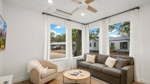 A living room with a sofa, armchair, coffee table, and large windows with curtains. - Guadalupe Cabins | 3 Cottages | Sleeps 17-23 (New Braunfels)