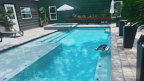 A rectangular swimming pool with a built-in hot tub, surrounded by a wooden deck and a green house in the background. - Purple Rain/Pop Culture 2bd 2bth Luxury Pool Hot Tub (Lutz)