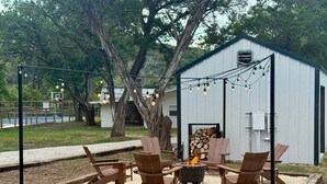 An outdoor seating area with wooden chairs and a fire pit, surrounded by a metal structure. - Waltz On In | 1BR King | Pool + River | Pets Welcome (New Braunfels)