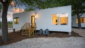 A cabin with a wooden staircase, a small porch with two chairs, and a window with curtains. - Waltz On In | 1BR King | Pool + River | Pets Welcome (New Braunfels)